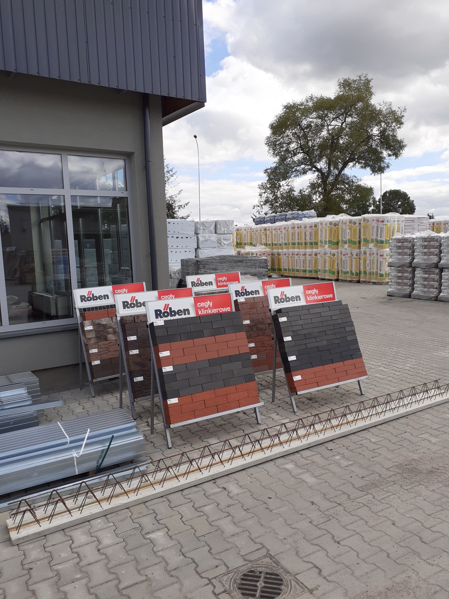 Ekspozycja cegieł klinkierowych marki Roben na zewnątrz budynku, w tle zapasy materiałów budowlanych i drzewo.
