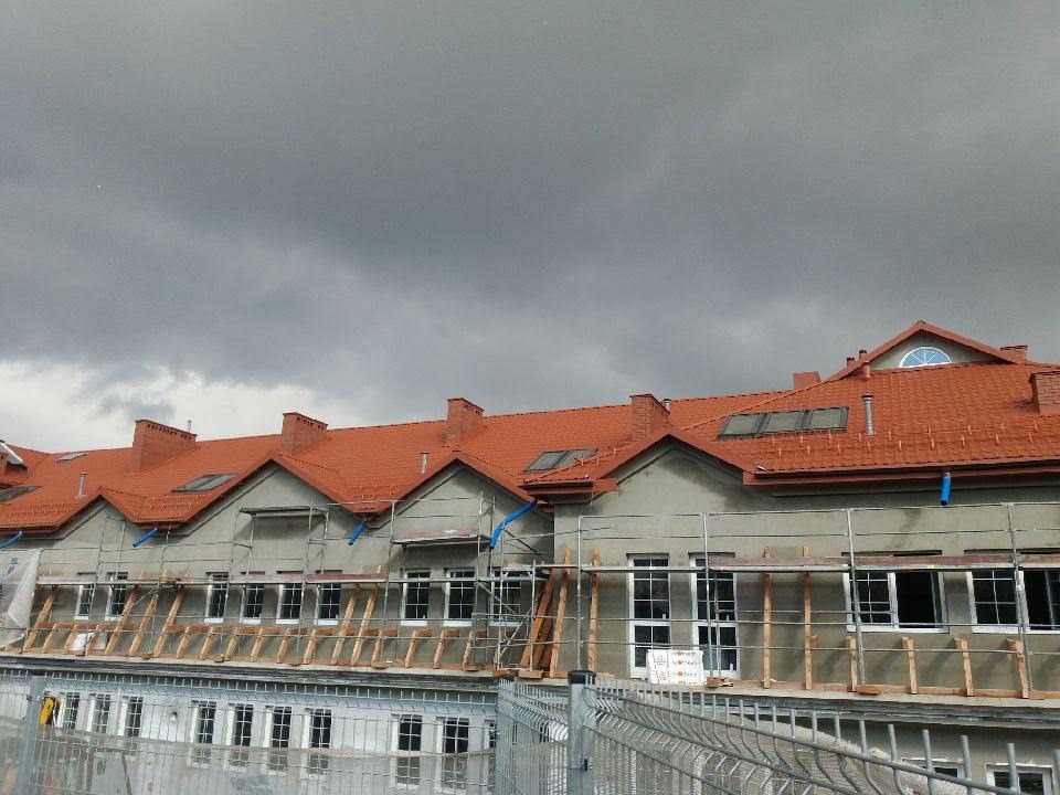 Budynek wielorodzinny w trakcie prac budowlanych, widoczne rusztowania, nowa dachówka w kolorze ceglastym, okna dachowe i kominy, na tle pochmurnego nieba.