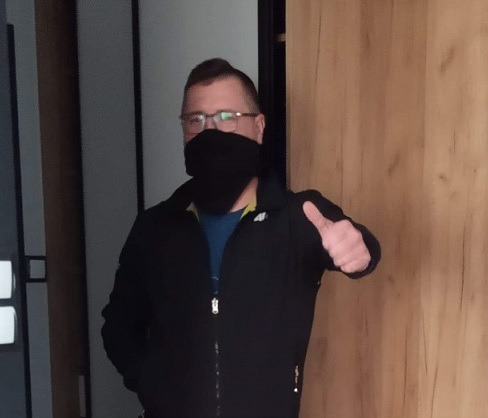 Mężczyzna w okularach i czarnej masce na twarzy, ubrany w czarną bluzę, pokazuje kciuk w górę na tle drewnianej ściany i szarych drzwi.