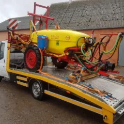 Żółty opryskiwacz rolniczy z czerwonymi elementami umieszczony na platformie lawety, zabezpieczony pasami transportowymi. W tle budynek gospodarczy z szarą bramą.