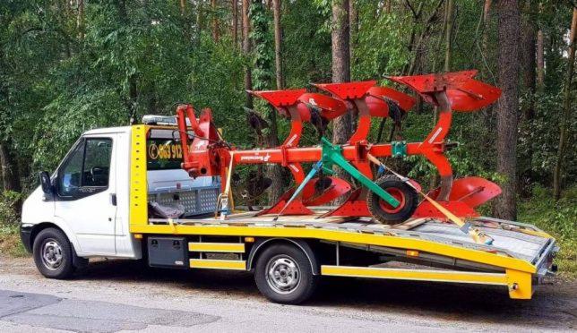 Biała laweta z żółtymi elementami przewozi czerwony pług rolniczy na tle drzew