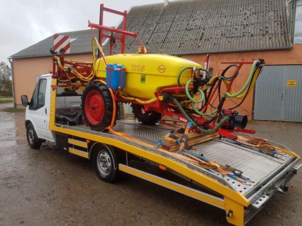 Żółty opryskiwacz rolniczy z czerwonymi elementami umieszczony na platformie lawety, zabezpieczony pasami transportowymi. W tle budynek gospodarczy z szarą bramą.