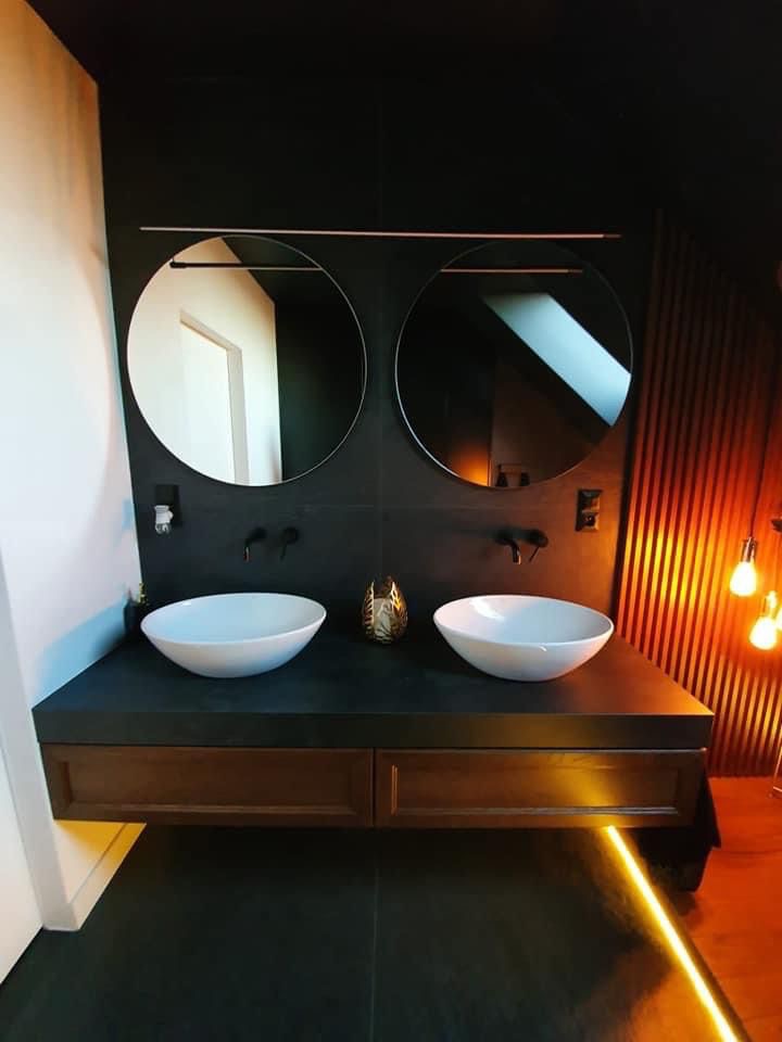 Nowoczesna łazienka z dwoma okrągłymi lustrami nad umywalkami nablatowymi. Ciemna kolorystyka, drewniane elementy i oświetlenie LED pod szafką.