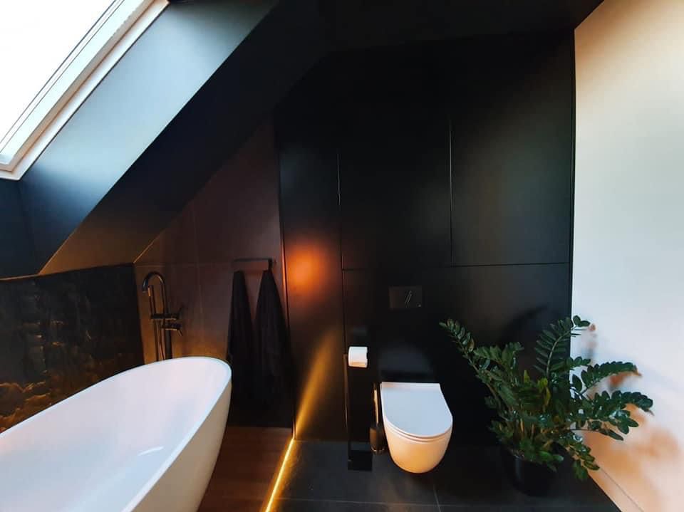 Nowoczesna łazienka na poddaszu w czerni i bieli. Wolnostojąca wanna, podświetlenie LED przy podłodze i roślina doniczkowa tworzą spójną, elegancką przestrzeń.