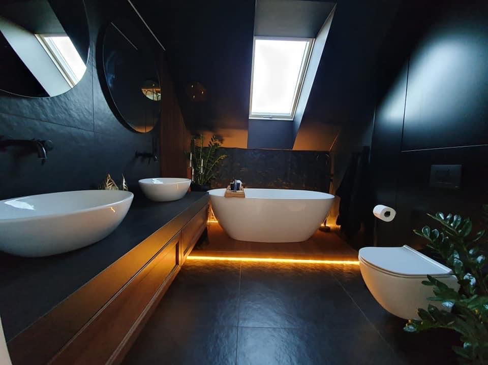 Nowoczesna łazienka na poddaszu z czarnymi płytkami, wolnostojącą wanną, dwiema umywalkami i podświetleniem LED pod szafką. Okrągłe lustra i roślina doniczkowa.