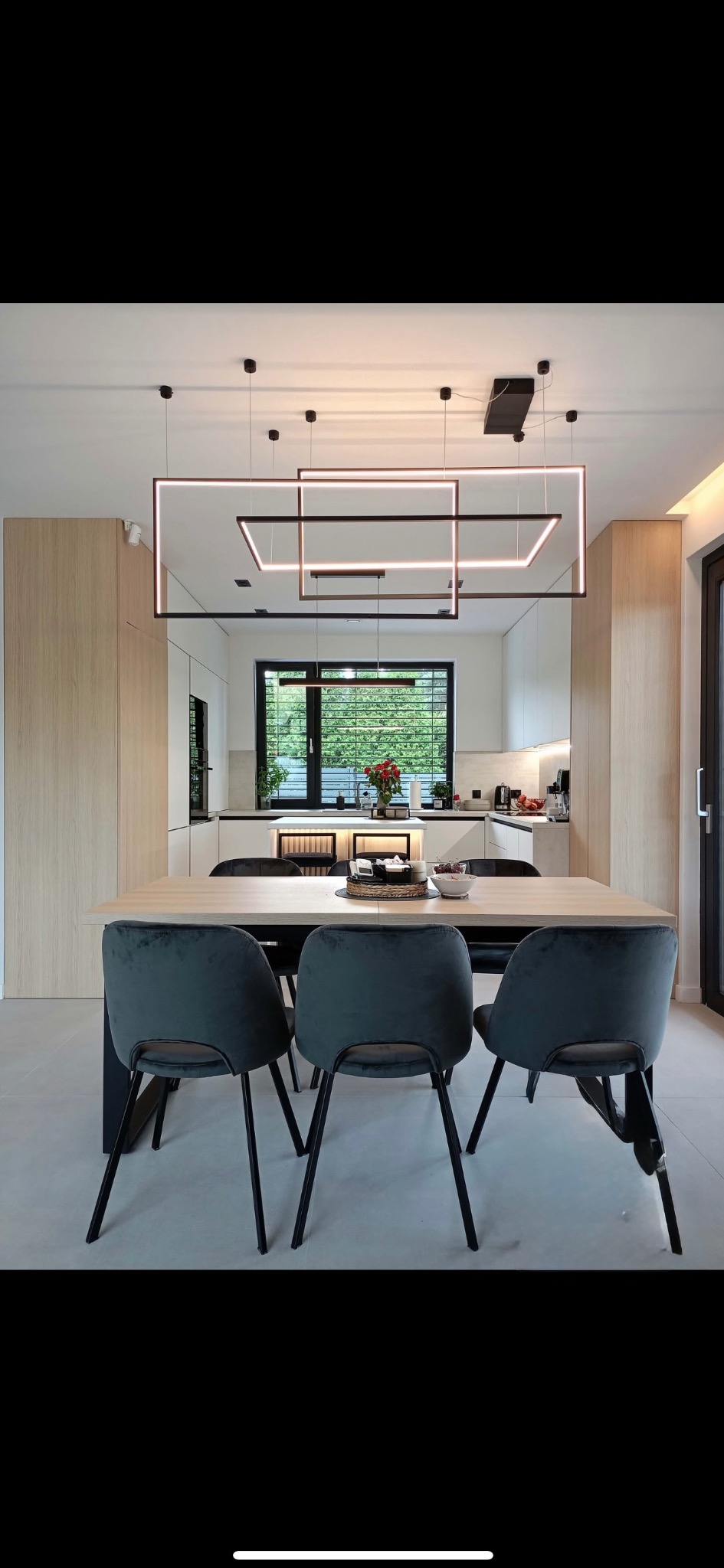 Jasna jadalnia z nowoczesną lampą sufitową z podświetlanych kwadratów, widok na kuchnię z oknem i zielenią. Minimalistyczny design wnętrza.