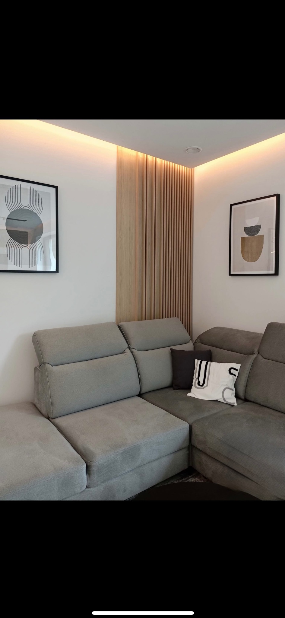 Szara, narożna sofa z poduszkami w nowoczesnym salonie. Na ścianach grafiki w czarnych ramkach, drewniane lamele i subtelne oświetlenie LED.