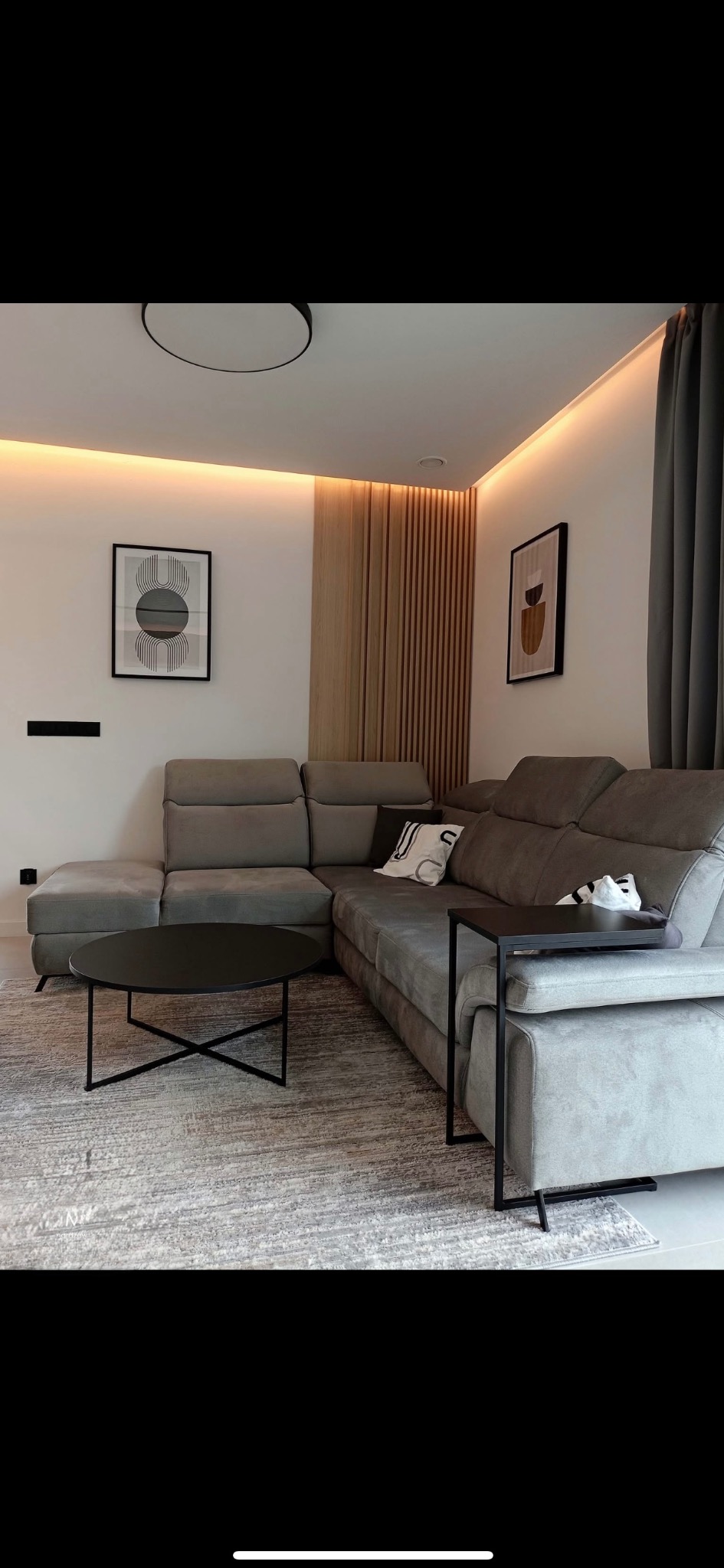 Stylowy salon z szarą narożną sofą, okrągłym stolikiem kawowym i drewnianymi lamelami na ścianie. Oświetlenie LED podkreśla nowoczesny design wnętrza.
