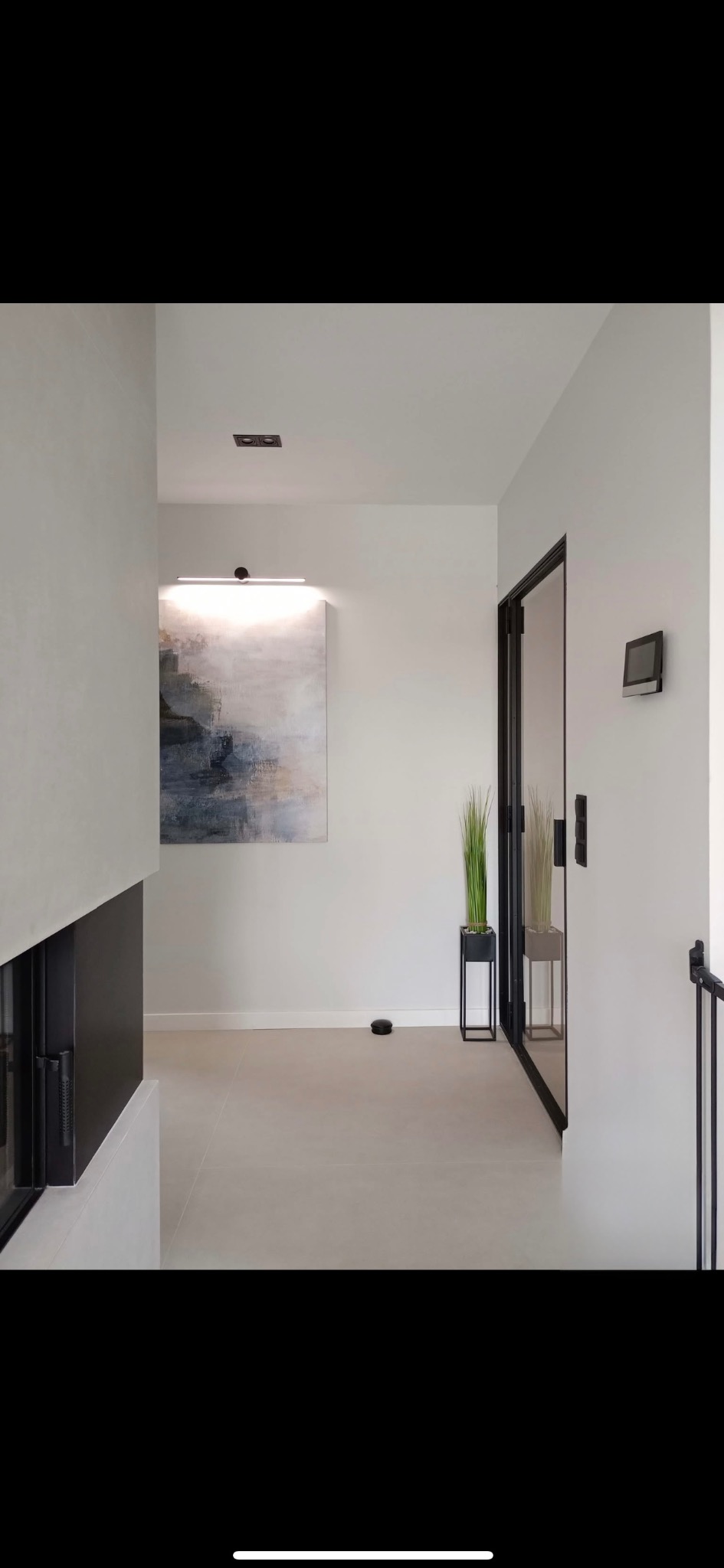 Minimalistyczny hol z szarą podłogą, ścianami w odcieniach bieli. Widoczny fragment kominka, obraz, roślina w czarnej donicy i czarne ramy drzwi.