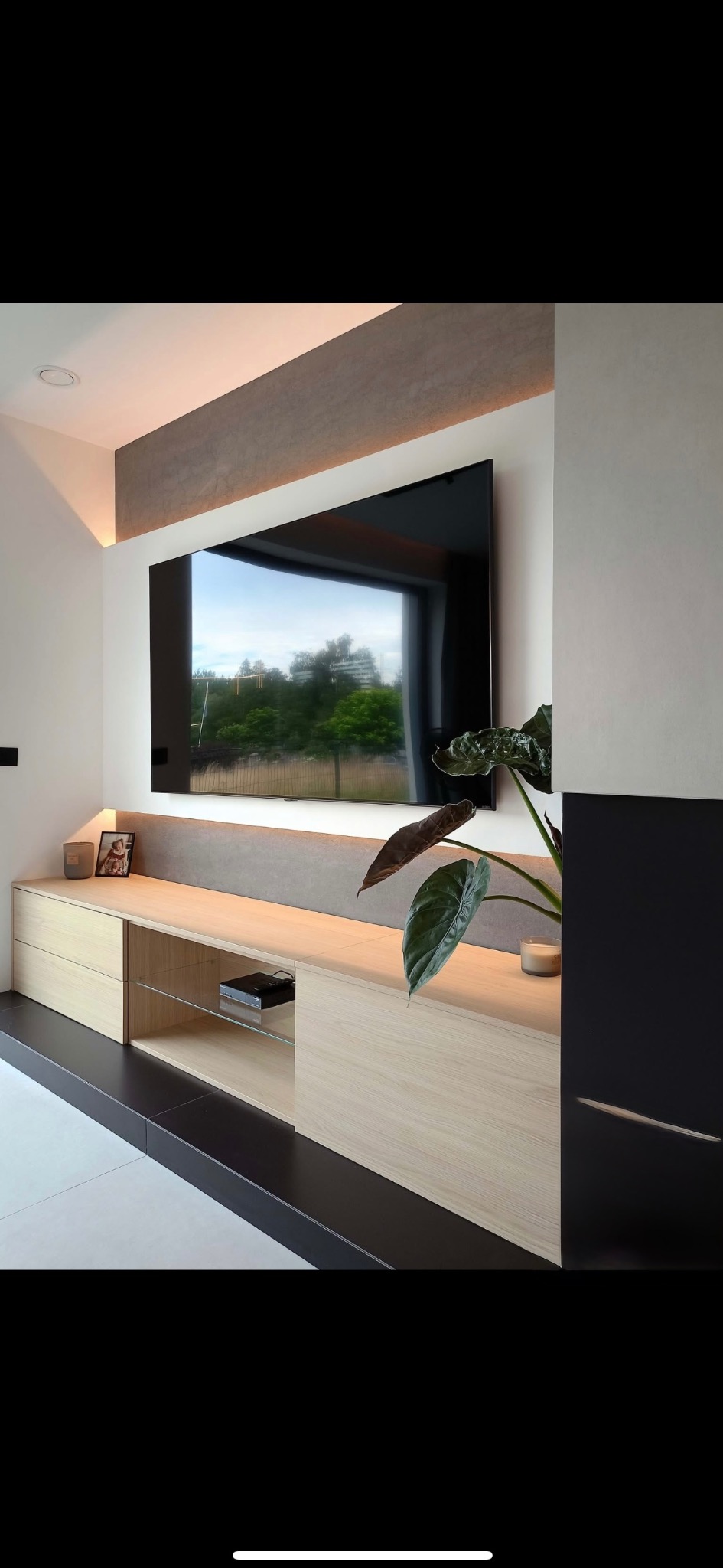 Nowoczesny salon z podświetlonym telewizorem i szafką RTV z jasnego drewna. Minimalistyczny design, roślina doniczkowa dodaje naturalnego akcentu. Oświetlenie LED podkreśla detale.