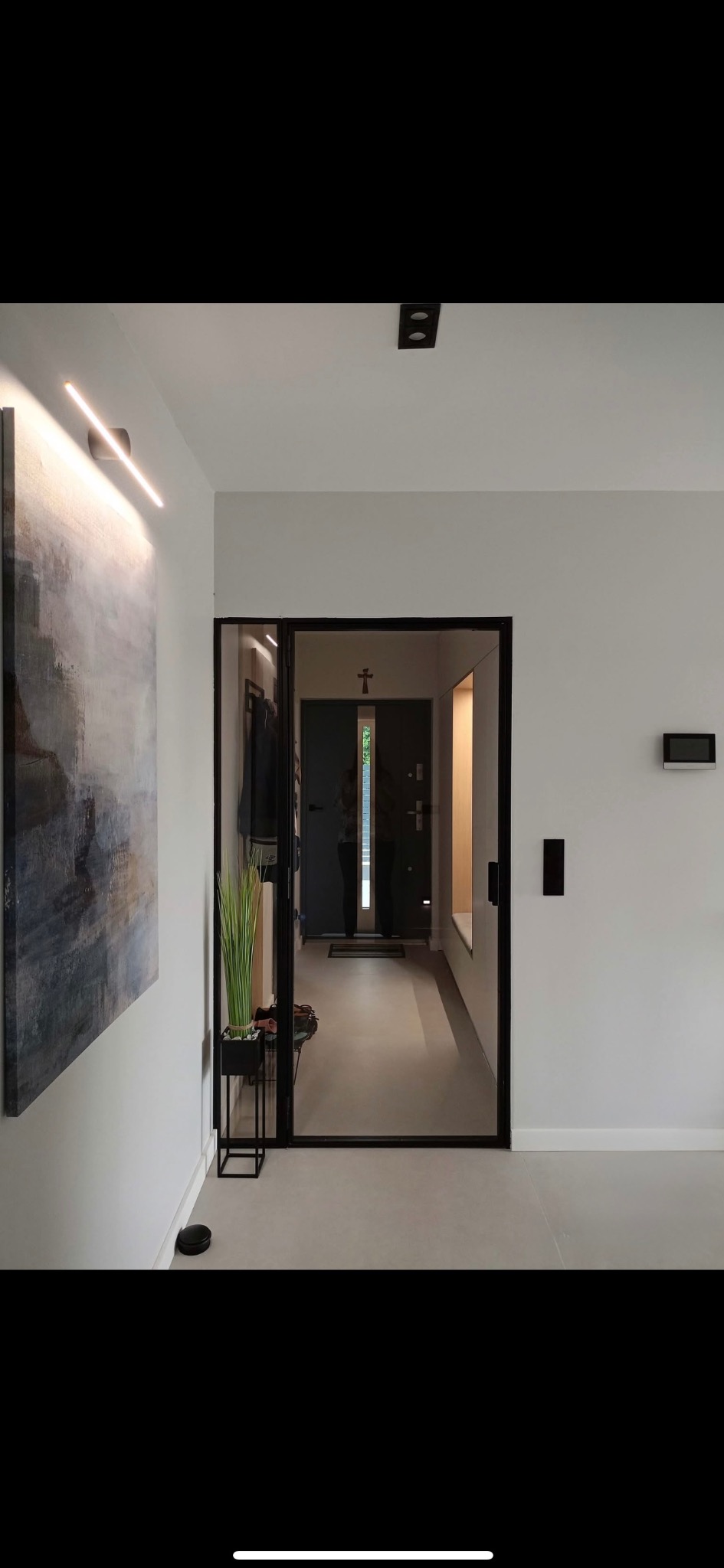 Nowoczesny przedpokój z przeszklonymi drzwiami w czarnej ramie. Minimalistyczny design, szara podłoga, abstrakcyjny obraz i oświetlenie LED.