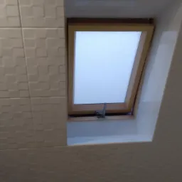 Uchylone okno dachowe z drewnianą ramą, zamontowane w łazience wyłożonej białymi, kwadratowymi płytkami z delikatnym wzorem 3D.