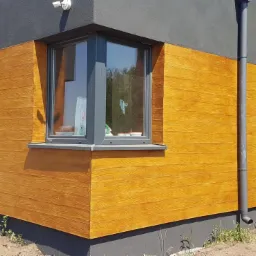 Fragment domu z elewacją w kolorze drewna, szare okna i ściany, widoczny narożnik budynku i rynna spustowa.