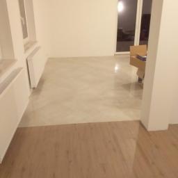 Usługi Remontowo-Budowlane Piotr Goworek - Wykończone wnętrze z podłogą częściowo pokrytą jasnobeżową żywicą, a częściowo panelami drewnopodobnymi; widoczne białe ściany, grzejniki oraz okno balkonowe.