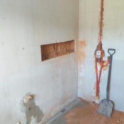 Usługi Remontowo-Budowlane Piotr Goworek - Ściana w trakcie prac przygotowawczych: wycięty otwór w murze, bruzdy pod instalację elektryczną z widocznymi przewodami w czerwonej osłonie, oraz łopata oparta o ścianę.