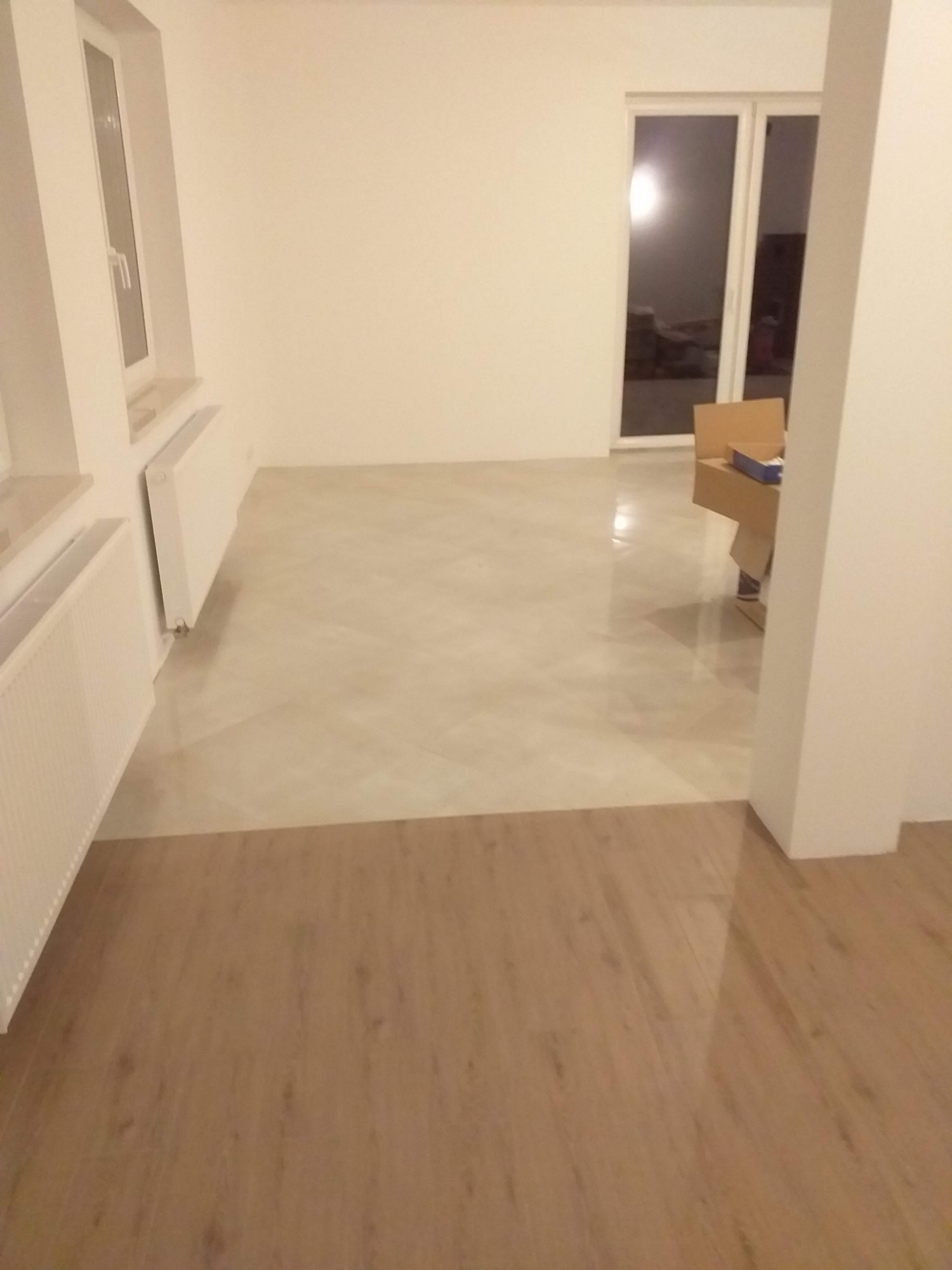 Wykończone wnętrze z podłogą częściowo pokrytą jasnobeżową żywicą, a częściowo panelami drewnopodobnymi; widoczne białe ściany, grzejniki oraz okno balkonowe.