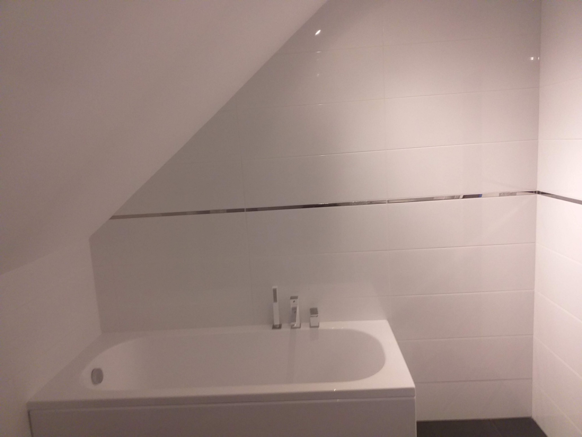 Biała wanna w łazience na poddaszu, wykończona białymi płytkami z poziomym srebrnym akcentem, widoczne baterie.