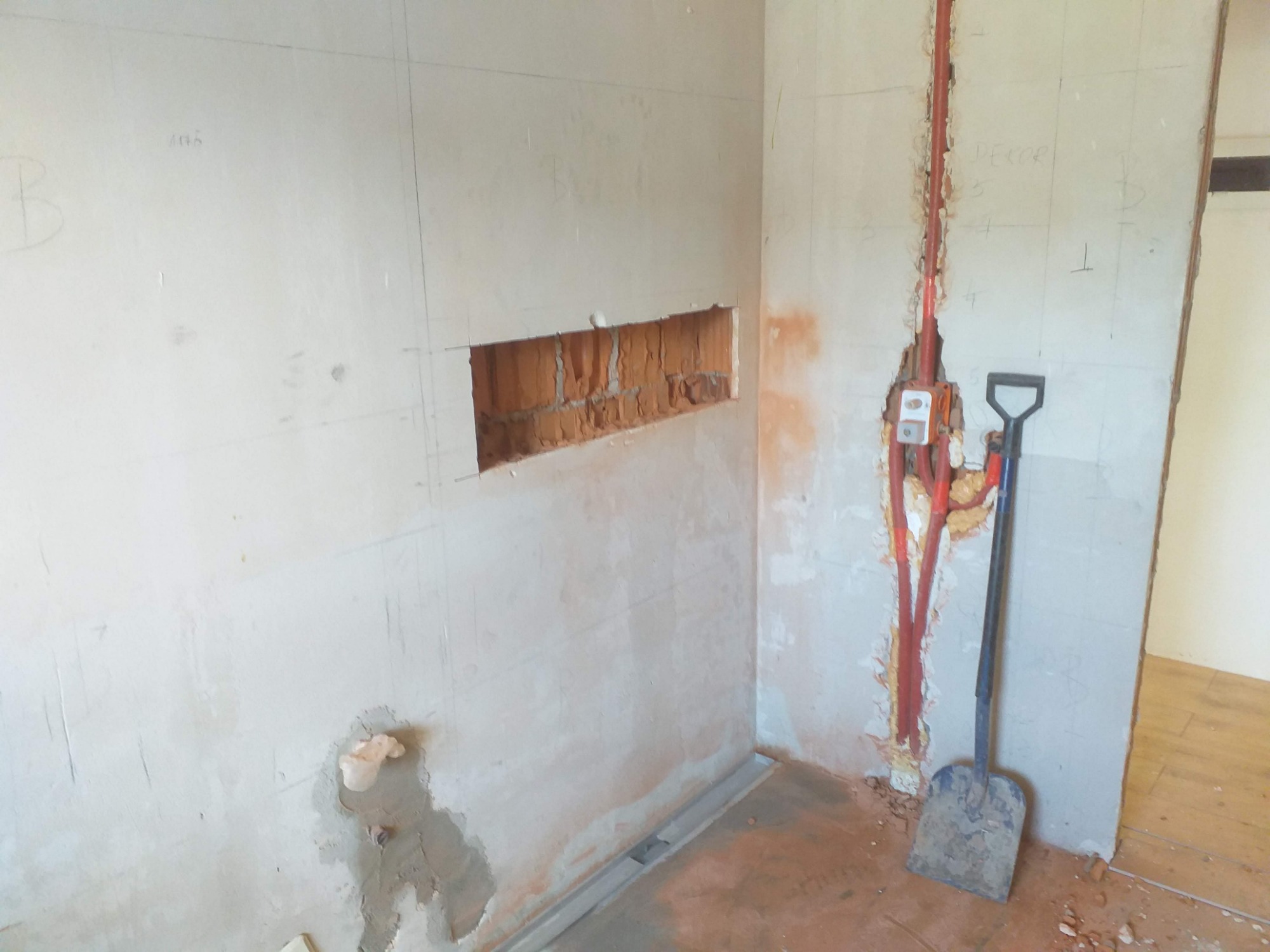 Ściana w trakcie prac przygotowawczych: wycięty otwór w murze, bruzdy pod instalację elektryczną z widocznymi przewodami w czerwonej osłonie, oraz łopata oparta o ścianę.