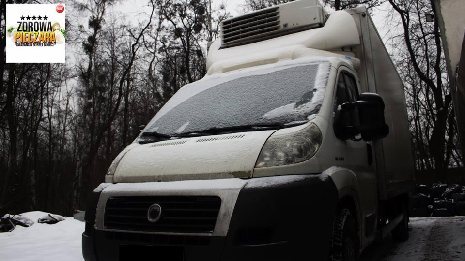 Biały samochód dostawczy z agregatem chłodniczym na dachu, lekko pokryty śniegiem, zaparkowany na tle zalesionego terenu w zimowej scenerii; widoczne logo firmy ZDROWIA PIECZARA w lewym górnym rogu.