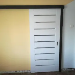 Białe drzwi przesuwne z poziomymi przeszkleniami zamontowane na ciemnej szynie, widok z boku na żółtą ścianę.