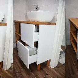 Łazienkowa szafka pod umywalkę z blatem z jasnego drewna, białe fronty szuflad, otwarte półki z boku, widoczna otwarta szuflada, brązowe panele podłogowe.