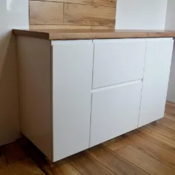 Biała szafka łazienkowa z drewnianym blatem, widoczny fragment ściany z białymi płytkami i drewnianą dekoracją, podłoga z paneli imitujących drewno, logo firmy na ścianie.