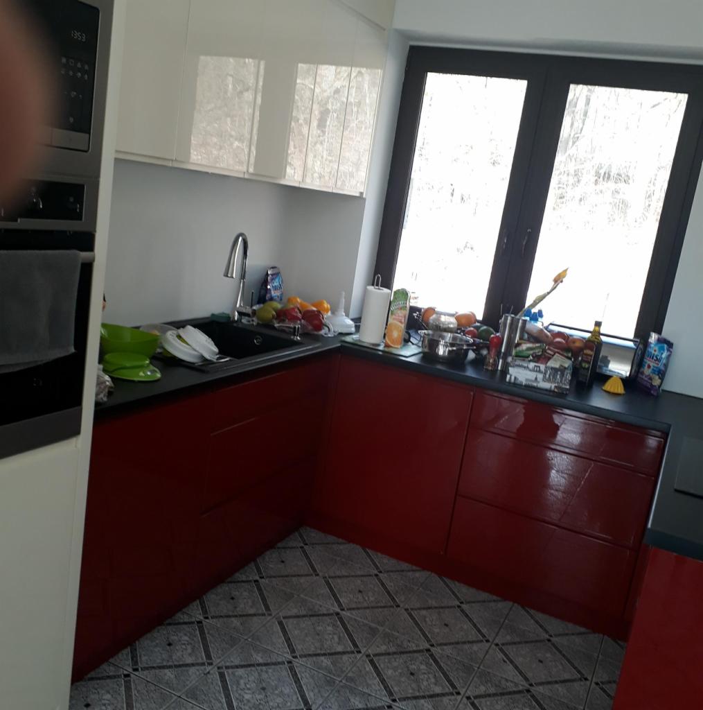 Nowoczesna kuchnia z czerwonymi szafkami dolnymi i białymi górnymi, czarnym blatem i oknem z widokiem na drzewa. Na blacie widoczne naczynia, owoce i akcesoria kuchenne.