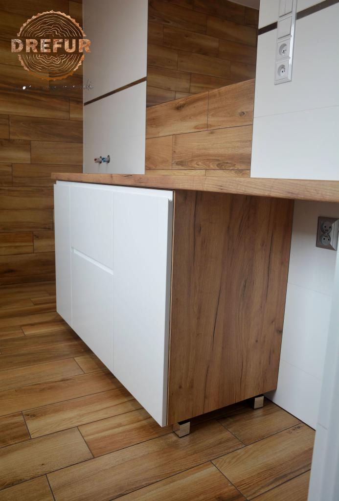Dolna szafka kuchenna z białymi frontami i drewnianym bokiem, z blatem w kolorze drewna, na metalowych nóżkach, dopasowana do drewnianej podłogi i ściany.