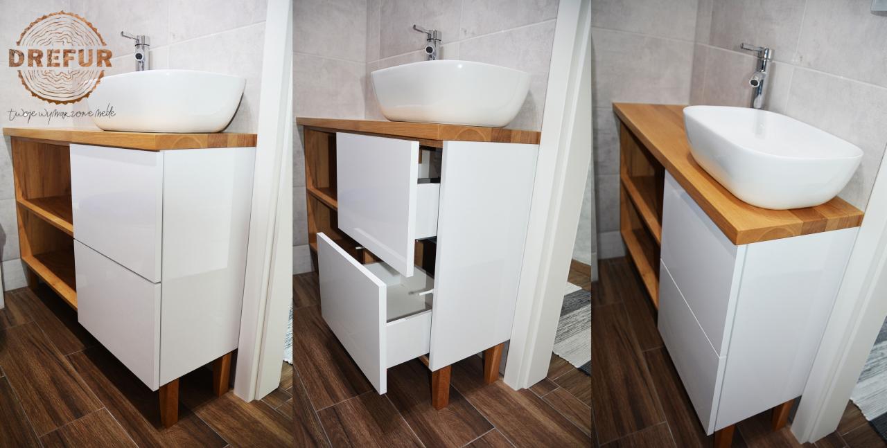 Łazienkowa szafka pod umywalkę z blatem z jasnego drewna, białe fronty szuflad, otwarte półki z boku, widoczna otwarta szuflada, brązowe panele podłogowe.