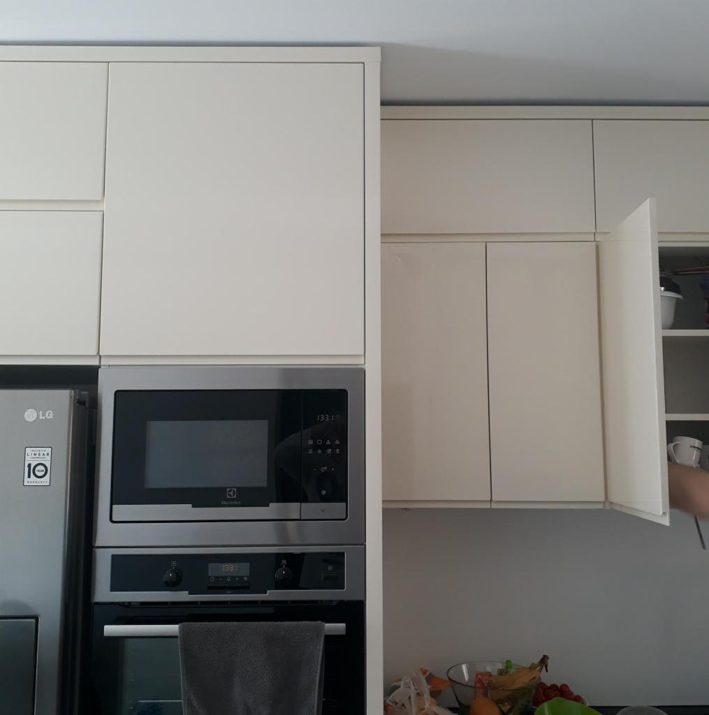 Jasne, lakierowane szafki kuchenne z wbudowanym piekarnikiem i mikrofalówką, otwarte drzwiczki szafki ukazujące zawartość półek, widoczna część blatu z owocami.