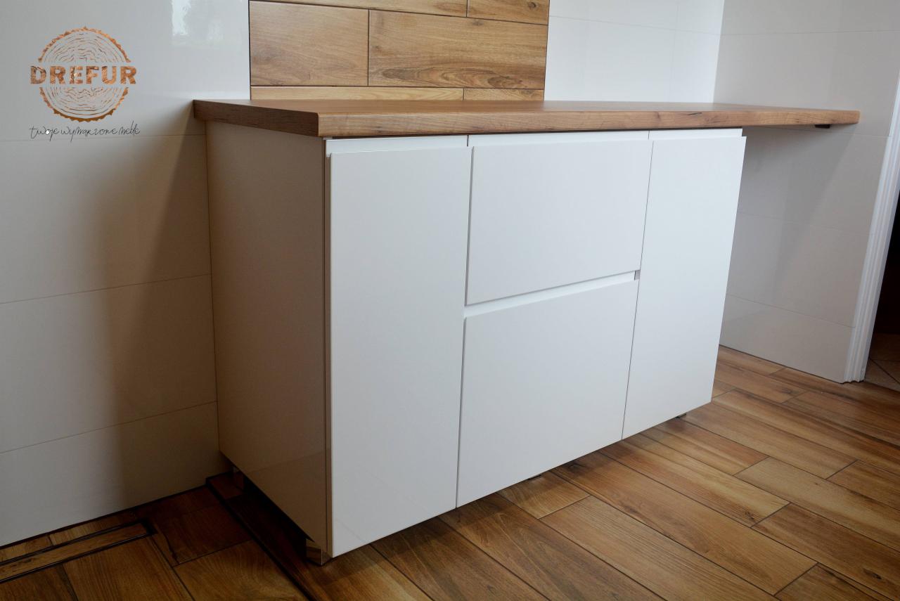 Biała szafka łazienkowa z drewnianym blatem, widoczny fragment ściany z białymi płytkami i drewnianą dekoracją, podłoga z paneli imitujących drewno, logo firmy na ścianie.