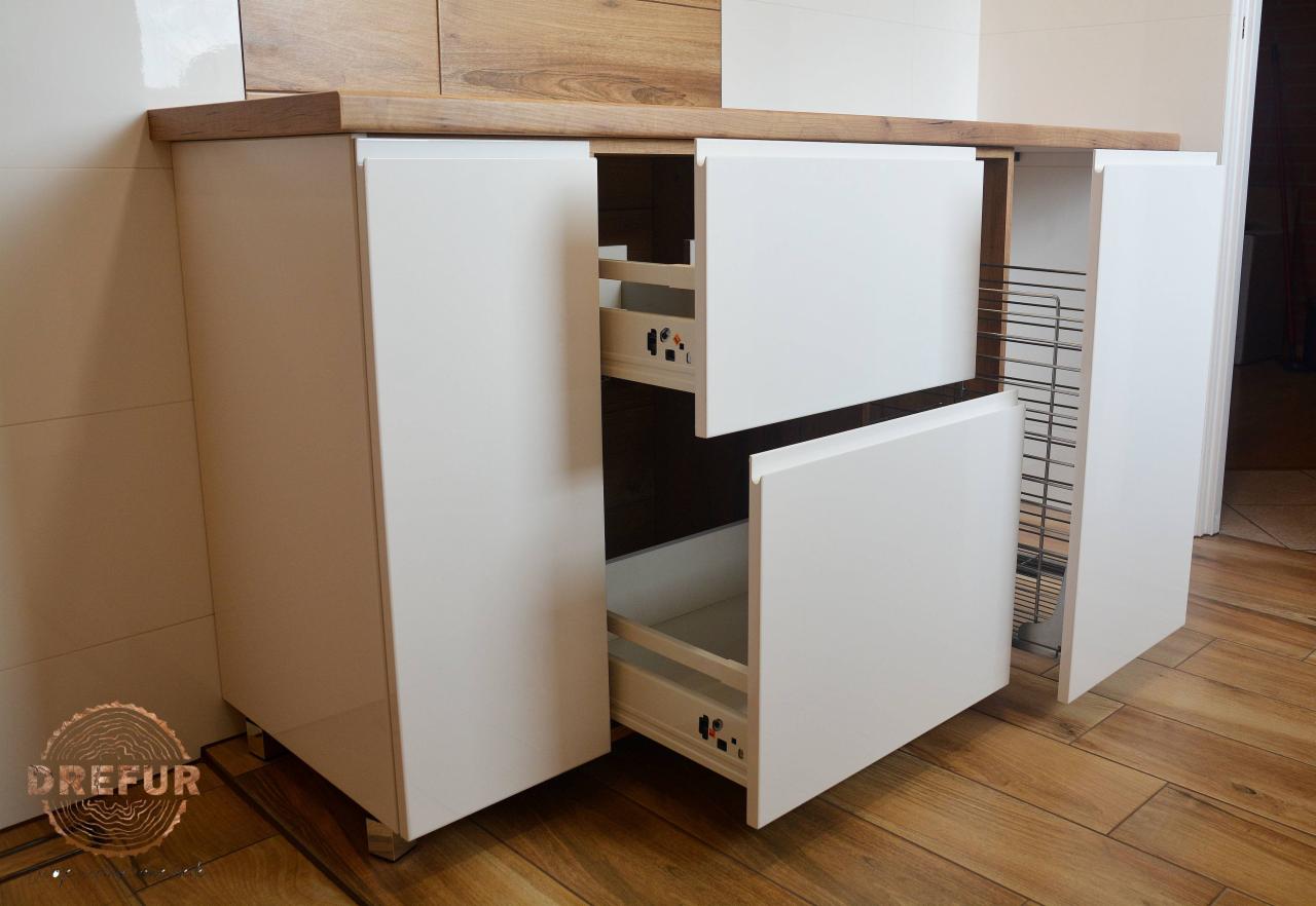 Biała szafka podblatowa z szufladami i koszem cargo, widoczne prowadnice i elementy montażowe, blat drewniany, podłoga drewnopodobna, fragment logo producenta w lewym dolnym rogu.