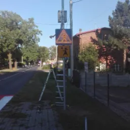 Montaż znaku drogowego zasilanego panelem słonecznym przy przejściu dla pieszych, drabina oparta o słup, widoczne narzędzia montażowe.