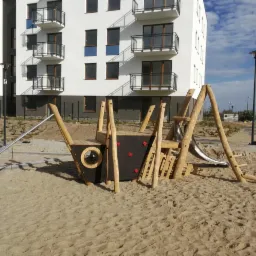 Drewniany plac zabaw w kształcie statku z metalową zjeżdżalnią na piaszczystym terenie, na tle nowego bloku mieszkalnego z balkonami.