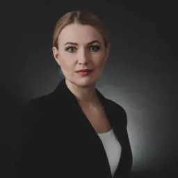 Portret kobiety w czarnej marynarce i białej bluzce na ciemnoszarym, rozmytym tle, z delikatnym uśmiechem i jasną karnacją.