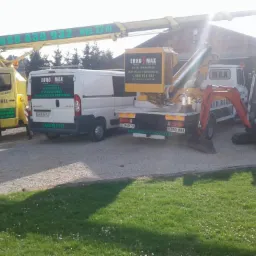 Żółty samochód z podnośnikiem koszowym, biały bus i minikoparka z logo firmy Euro Max na żwirowym podłożu przed budynkiem z cegły, w tle drzewa.