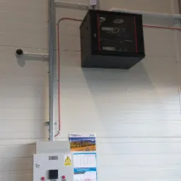 Szafa serwerowa zamontowana pod sufitem hali produkcyjnej, widoczne kamery monitoringu, skrzynka elektryczna z przyciskami i kalendarz na ścianie z blachy falistej.
