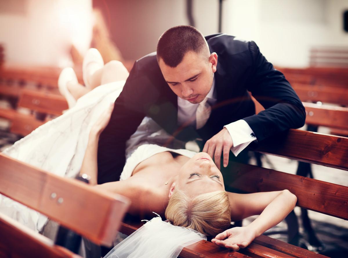 Panna młoda w sukni ślubnej leży na ławce w kościele, a pan młody w garniturze pochyla się nad nią, delikatnie dotykając jej twarzy.