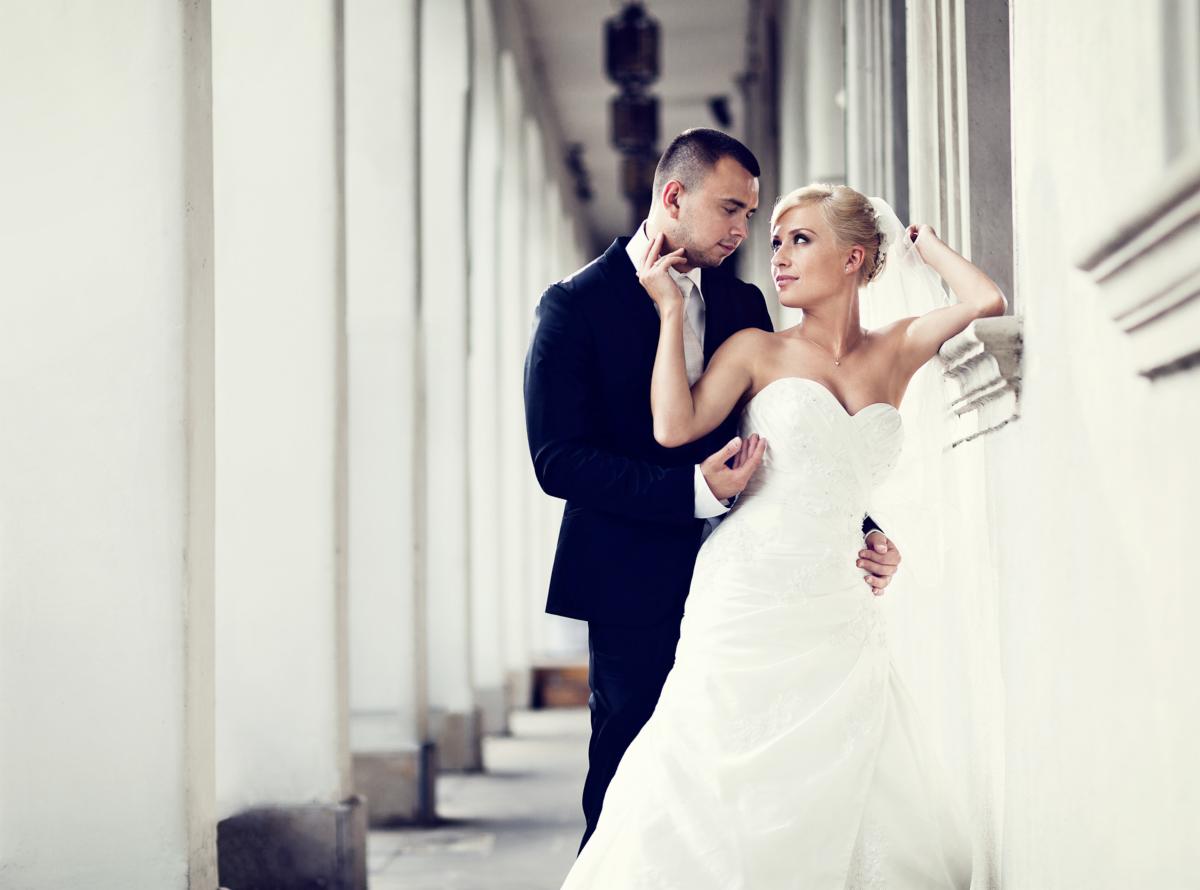 Para młoda: Panna młoda w białej sukni bez ramiączek, oparta o kolumnę, spogląda na pana młodego w czarnym garniturze, który delikatnie dotyka jej twarzy. W tle rząd białych kolumn.