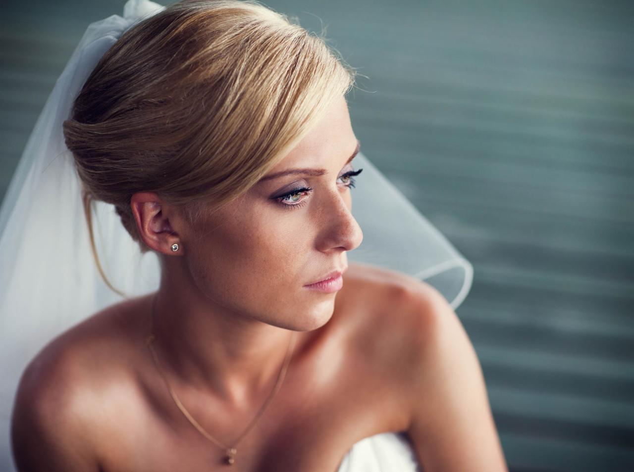 Portret panny młodej w welonie, z delikatnym makijażem i złotą biżuterią, spoglądającej w bok na tle rozmytego tła.