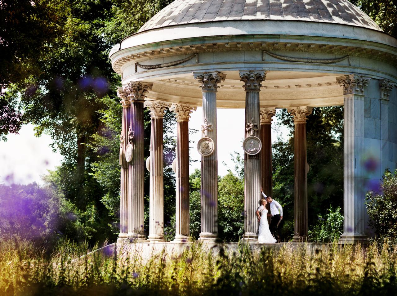 Para młoda w białej sukni i czarnym garniturze pozuje w klasycznej, okrągłej altanie z kolumnami ozdobionymi medalionami, otoczonej zielenią, w słoneczny dzień.