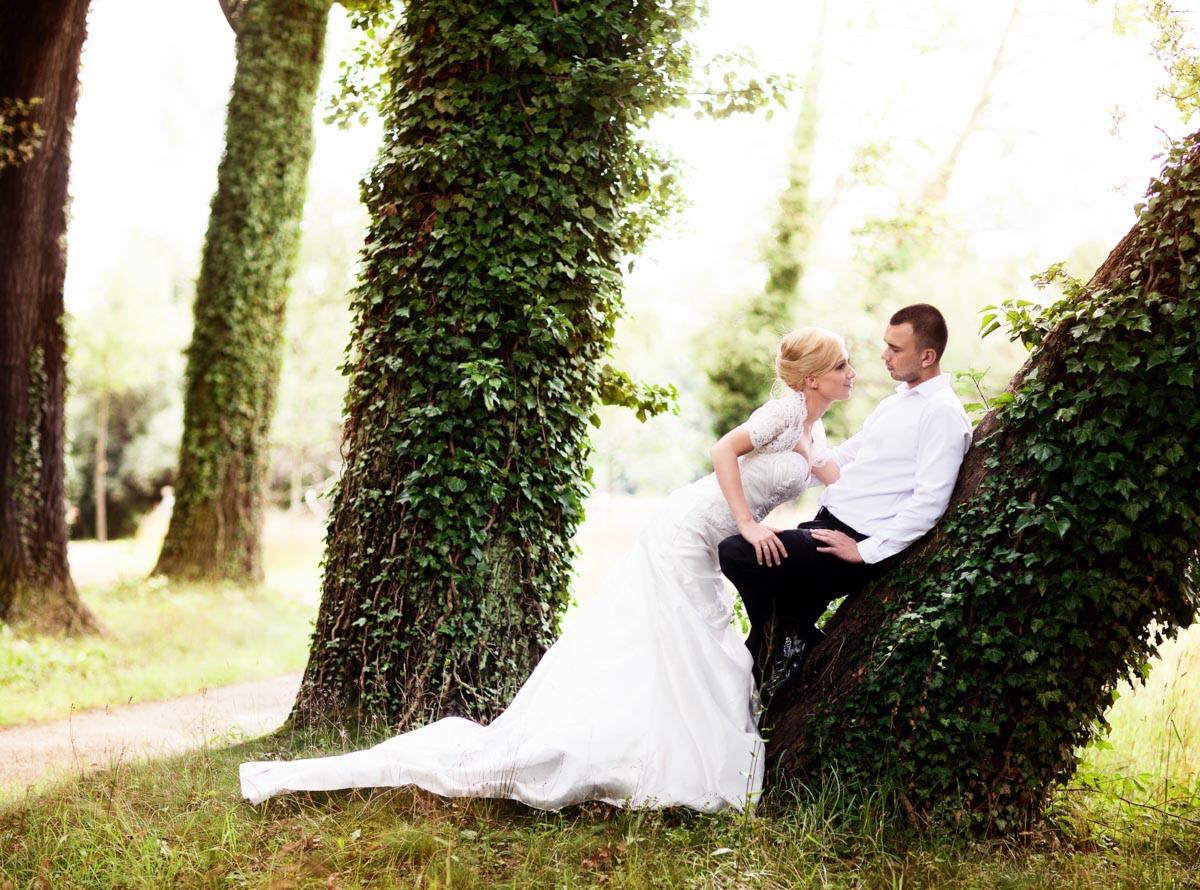 Para młoda w plenerze: Panna młoda w koronkowej sukni opiera się o drzewo obrośnięte bluszczem, spoglądając na siedzącego obok pana młodego w białej koszuli i czarnych spodniach.