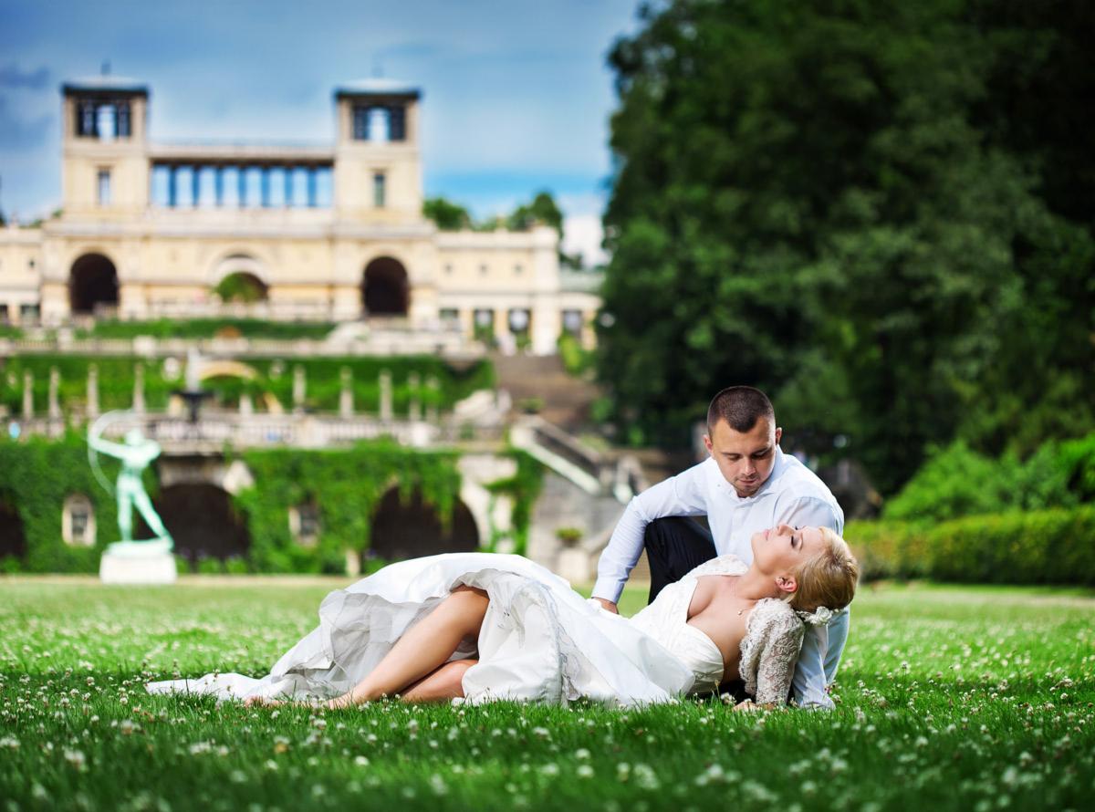Panna młoda w białej sukni leży na trawie, opierając głowę o kolana pana młodego. W tle park z budynkiem porośniętym bluszczem i rzeźbą łucznika.