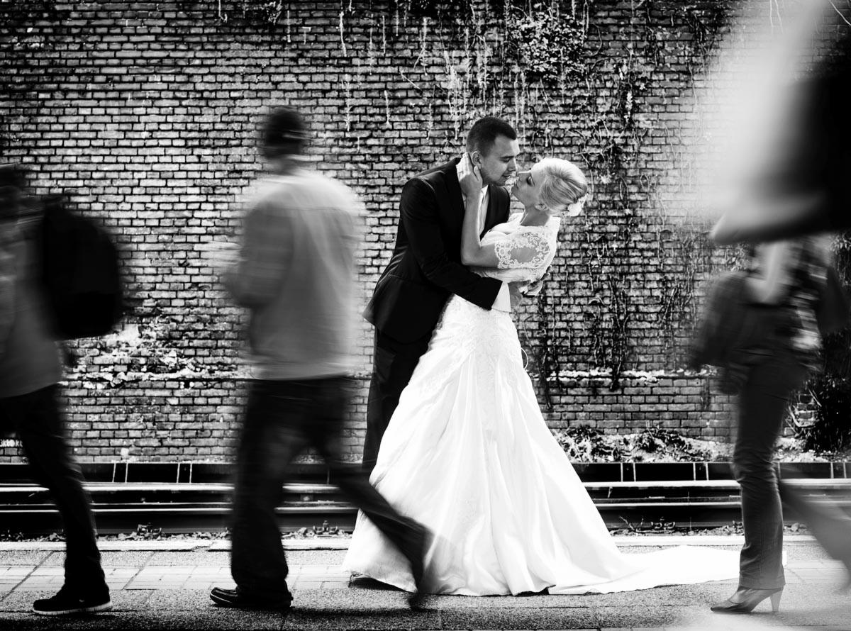 Czarno-białe zdjęcie całującej się pary młodej na tle ceglanej ściany z przechodzącymi, rozmytymi postaciami.