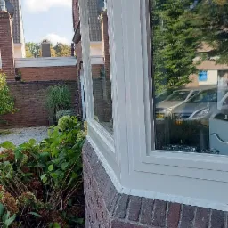 Malowanie ramy okiennej na biało, widoczna ceglana elewacja budynku i fragment ogrodu z krzewami.