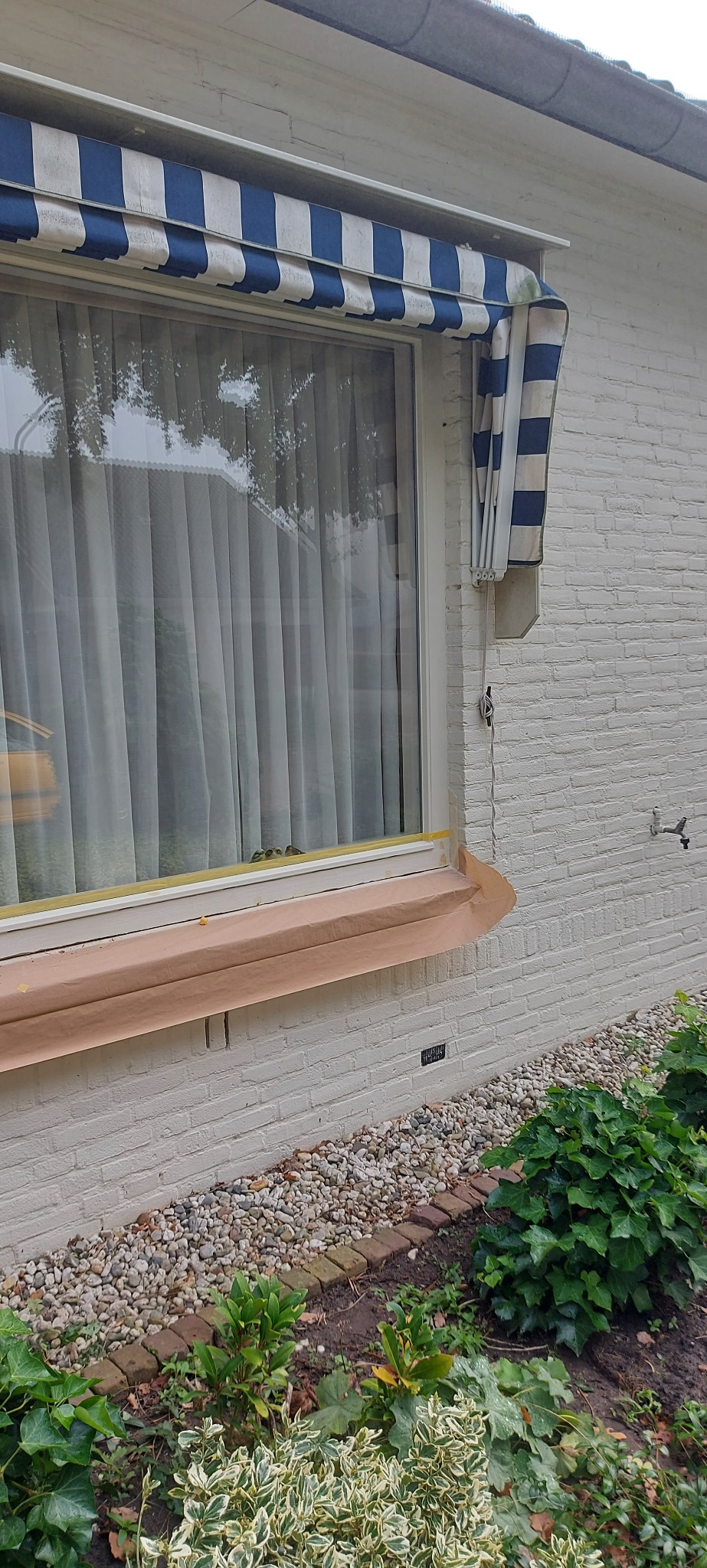 Okno zabezpieczone taśmą i papierem malarskim przed malowaniem elewacji, widoczna biała ceglana ściana budynku z niebiesko-białą markizą.