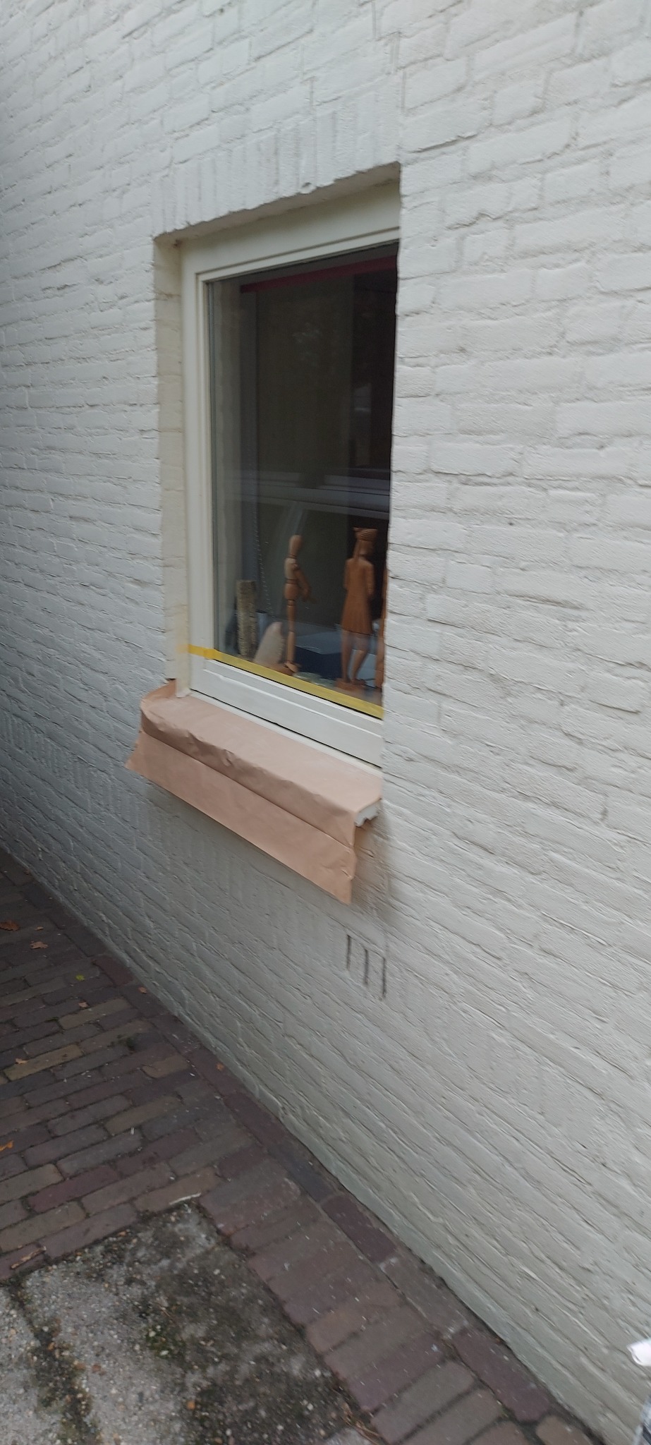 Biała ceglana elewacja z oknem zabezpieczonym papierem malarskim przed malowaniem, wewnątrz widoczne drewniane figurki.