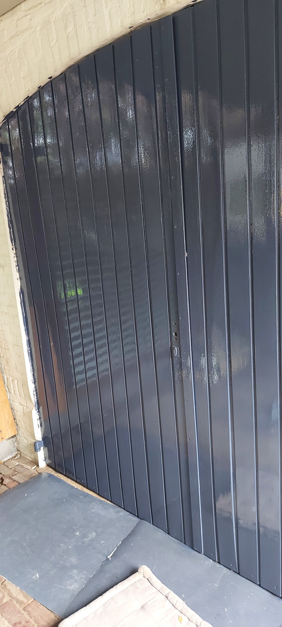Świeżo pomalowane, ciemnoszare, drewniane drzwi garażowe z widocznymi pionowymi panelami i zamkiem, z fragmentem muru z cegły w tle.