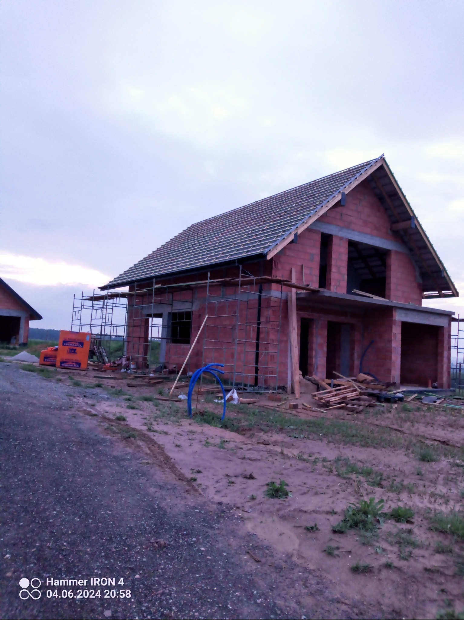 Surowy stan domu w trakcie budowy z czerwonej cegły, częściowo pokryty dachówką, z widocznymi rusztowaniami i materiałami budowlanymi na placu budowy.