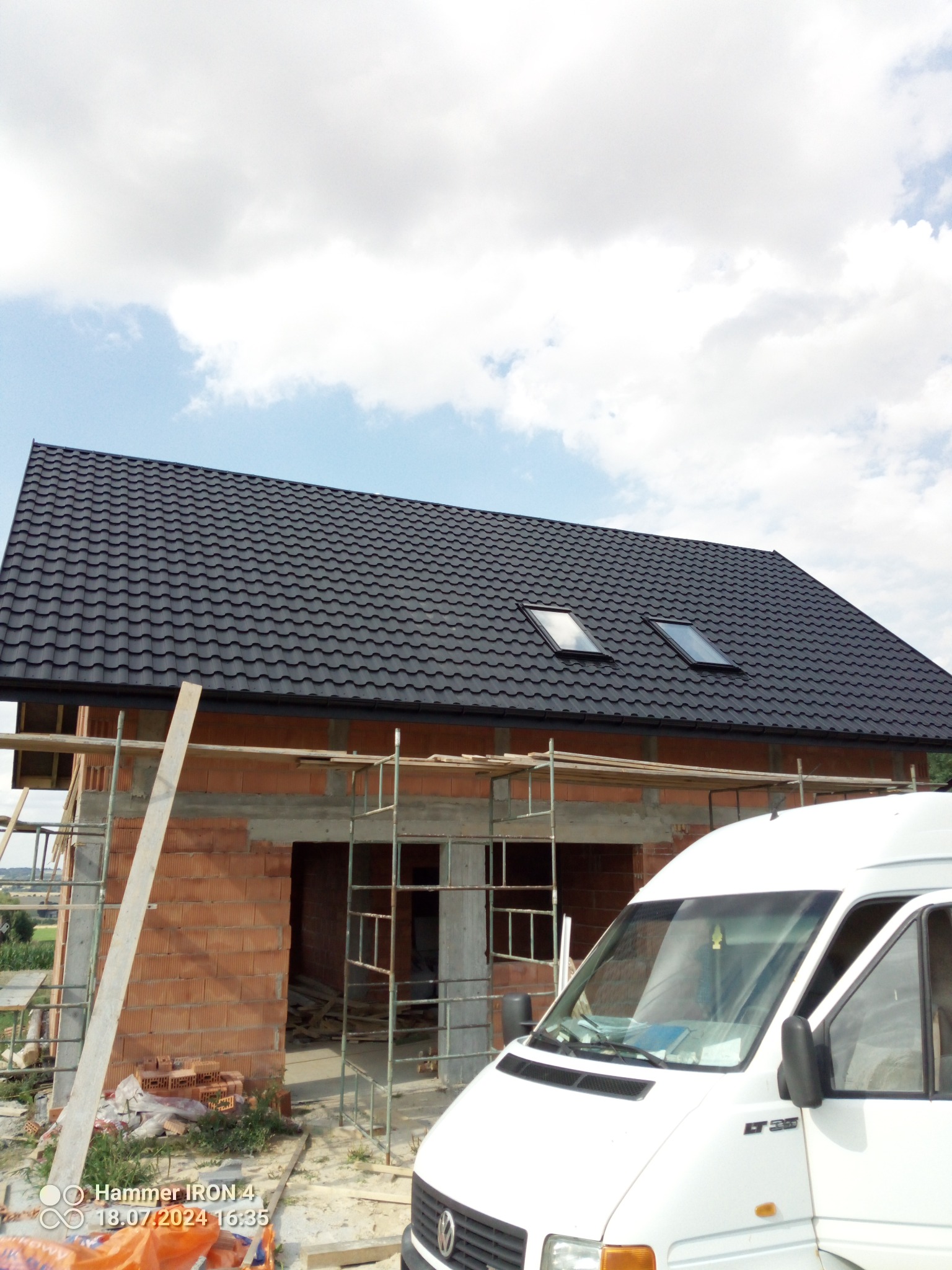 Budowa domu jednorodzinnego z ciemnym, metalowym dachem i dwoma oknami dachowymi, widok z poziomu ziemi, z przodu zaparkowany biały samochód dostawczy.